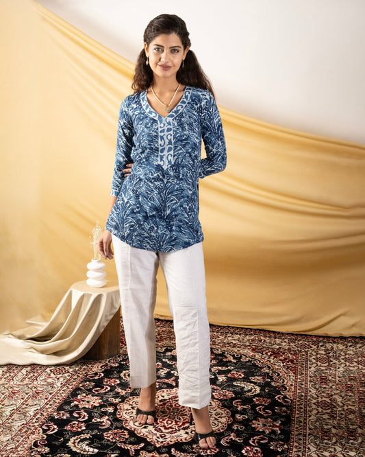 Woman wearing a blue patterned top and white pants standing on a decorative rug with a beige draped fabric in the background.